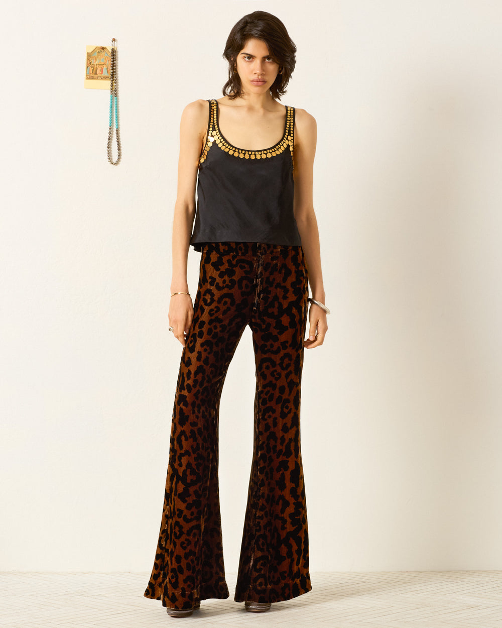 A person stands against a plain wall wearing the Fiona Gilded Coin Top, embellished with gold detail, and high-waisted, wide-leg, brown and black leopard print pants. A beaded necklace with golden metal coins hangs on the wall.