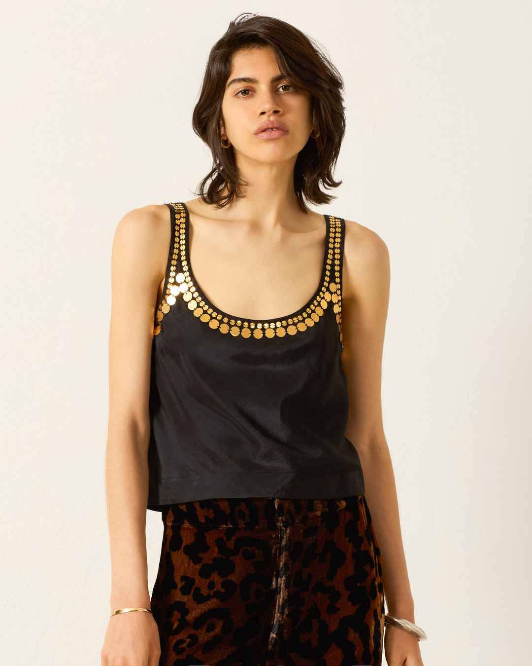 Wearing the Fiona Gilded Coin Top, featuring gold circular details at the neckline, paired with dark leopard print pants, the person stands against a plain white background.