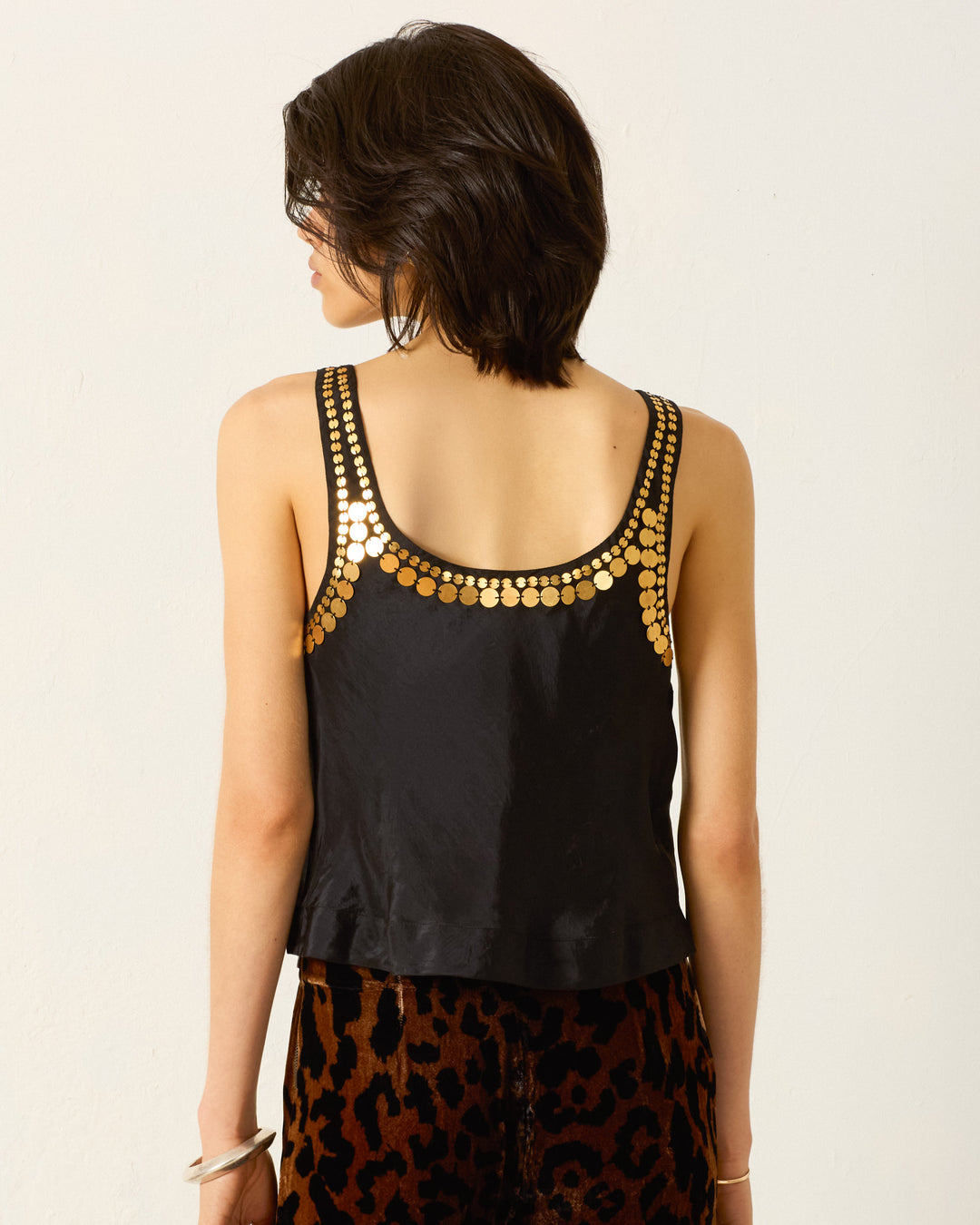 A person with short dark hair is shown from the back wearing the Fiona Gilded Coin Top, which features golden metal coins along the neckline, paired with leopard print pants.