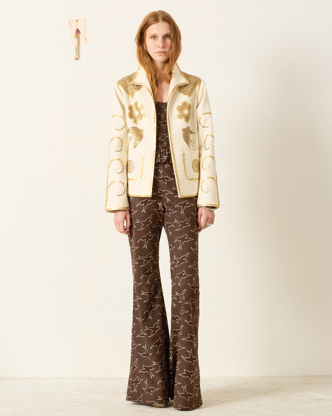 A woman stands against a plain wall wearing the Francis Pearl Nocturne Jacket over a brown patterned top and flared pants.