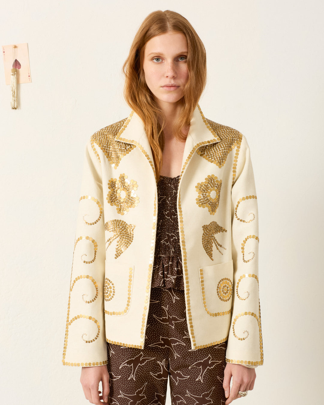 A woman with long reddish hair wears the Francis Pearl Nocturne Jacket, featuring gold sequin floral and swirl patterns over cream, paired with a brown patterned outfit, standing against a plain white wall.