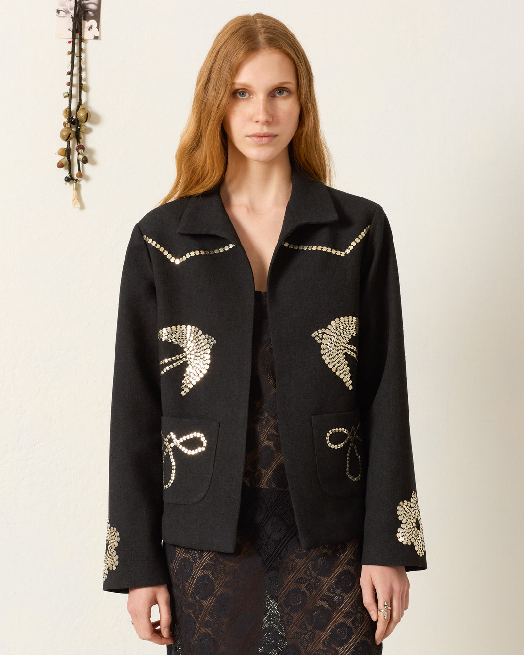 A woman with long, light brown hair wears the Francis Lovebird Coin Jacket, featuring hand-embellished gold designs, over a dark lace dress against a plain light background.