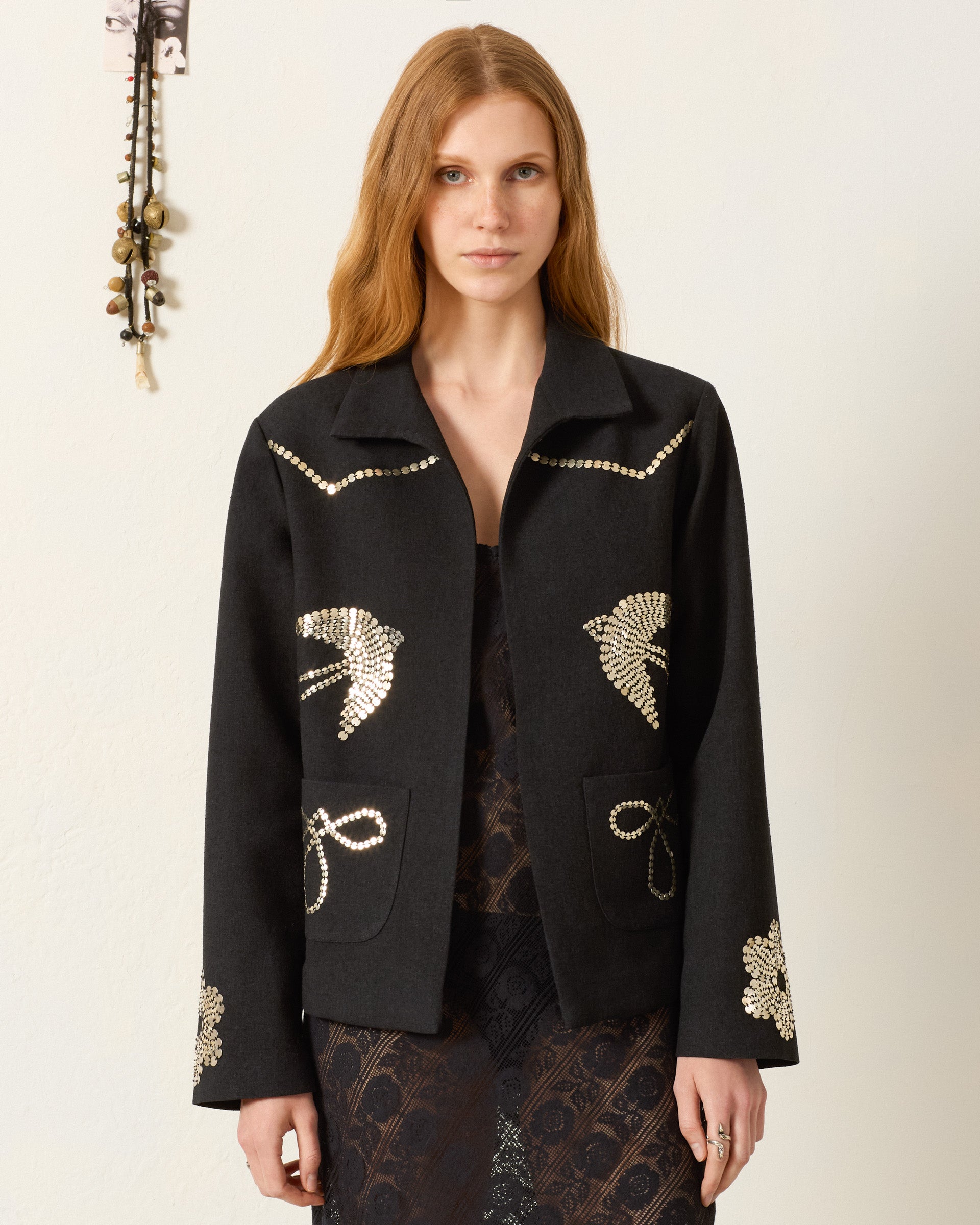 A woman with long, light brown hair wears the Francis Lovebird Coin Jacket, featuring hand-embellished gold designs, over a dark lace dress against a plain light background.