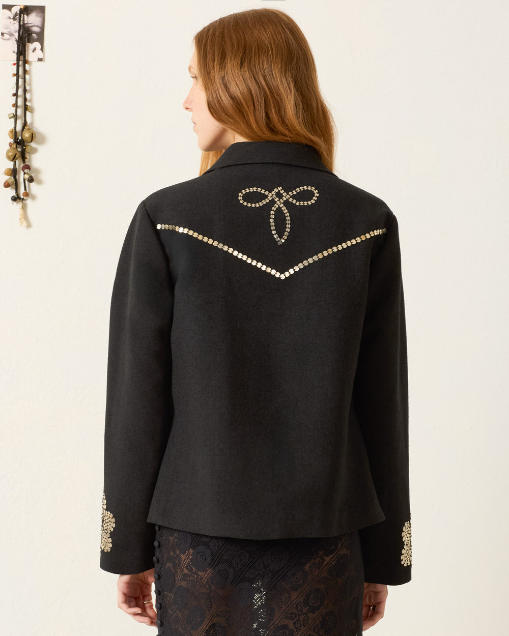 A person with long hair wears the Francis Lovebird Coin Jacket, featuring hand-embellished beading on the back and sleeves, while standing against a light-colored wall.