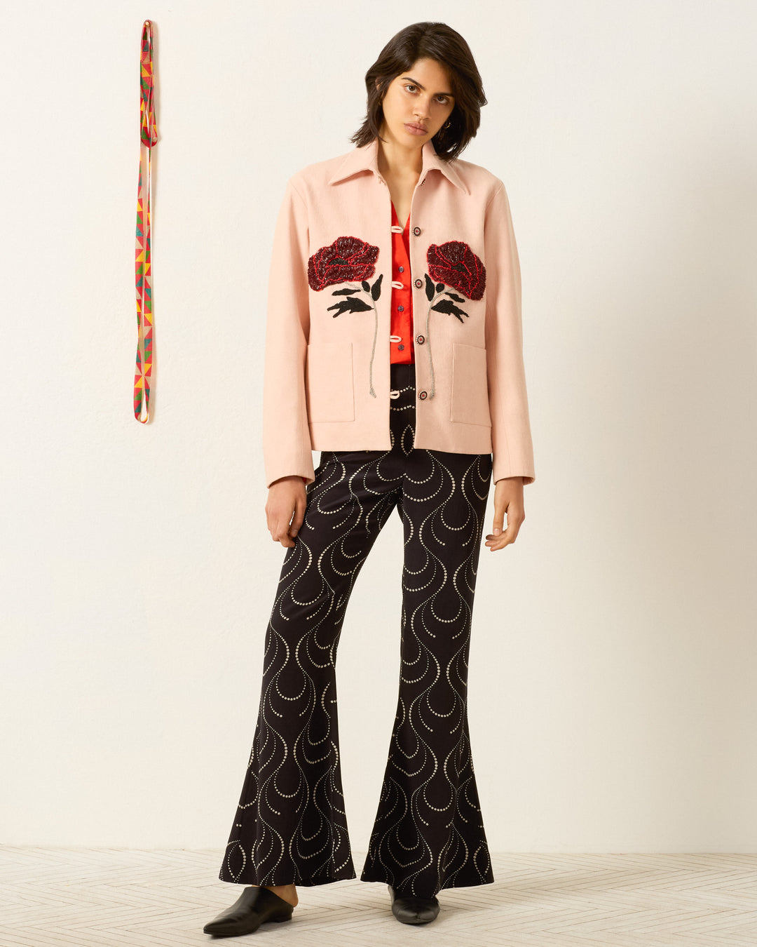 A person stands against a white wall wearing the Jude Ruby Poppy Jacket, a red shirt, black patterned flared pants, and black shoes. A colorful strap hangs on the wall.