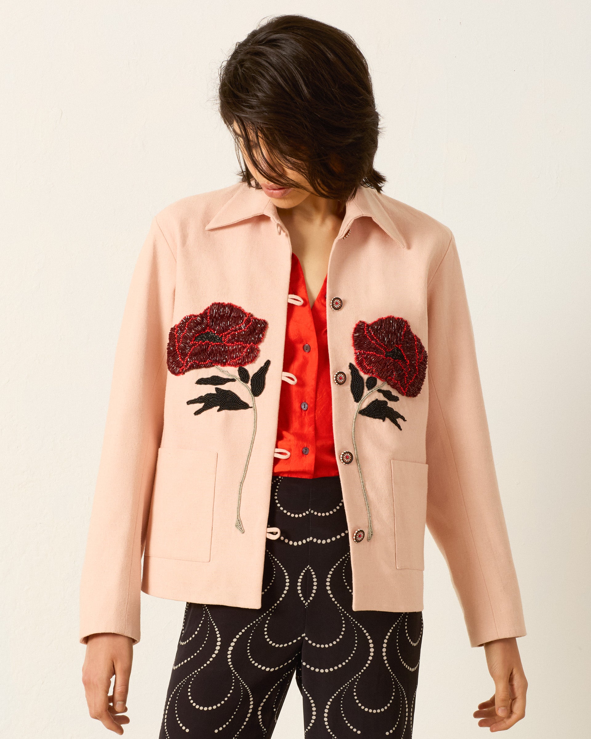Model wears the Jude Ruby Poppy Jacket, a light pink embellished piece with large red floral designs, layered over a red shirt and black pants featuring white swirl patterns, set against a plain light background.