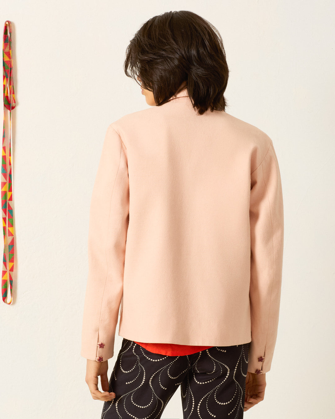 A person with short dark hair is seen from the back indoors, wearing the Jude Ruby Poppy Jacket and patterned black pants, standing next to a colorful hanging strap.