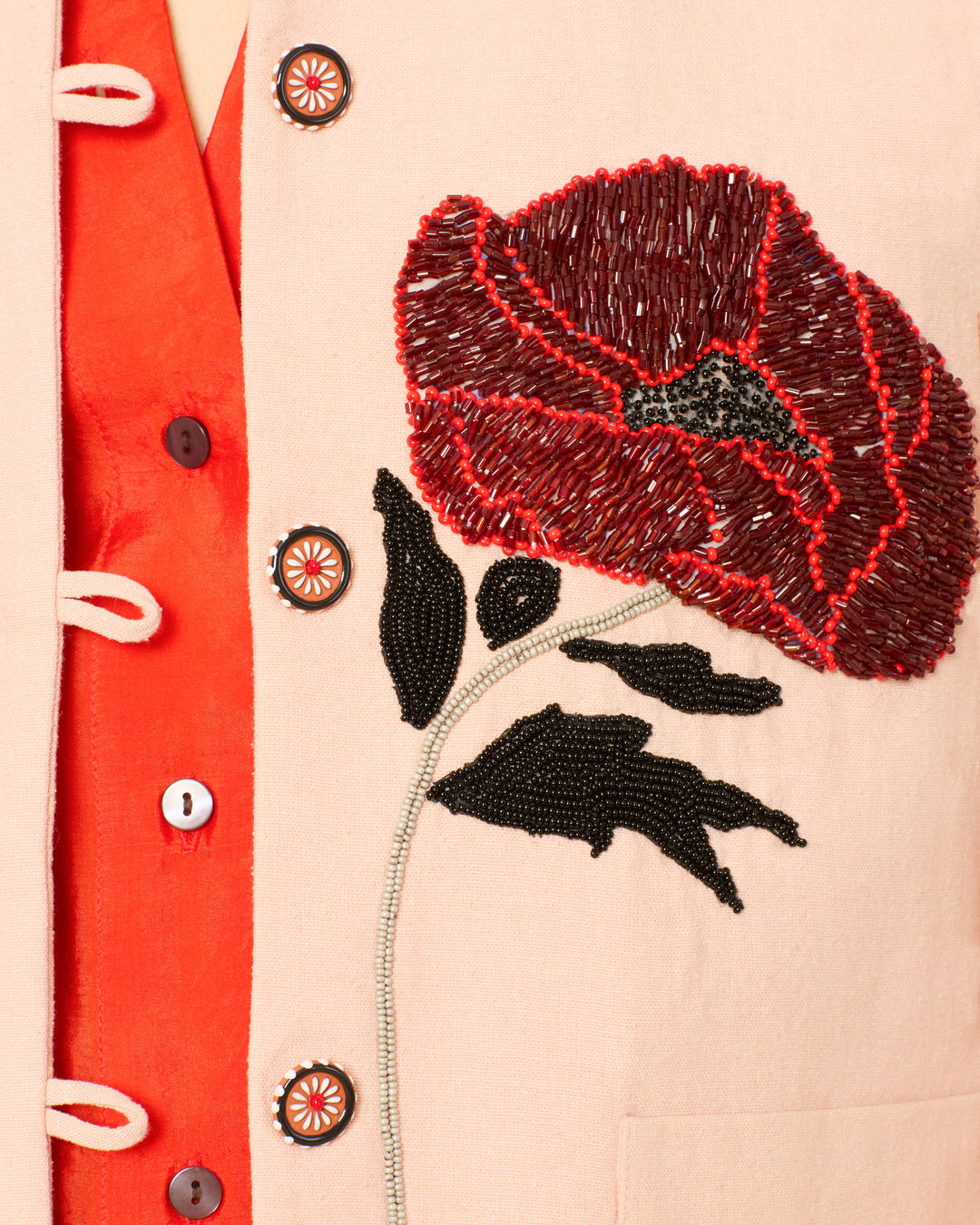 Close-up of the Jude Ruby Poppy Jacket, a hand-beaded beige textured silk piece featuring bold red poppy floral designs with black leaves, worn over an orange shirt with dark buttons and white loop closures.