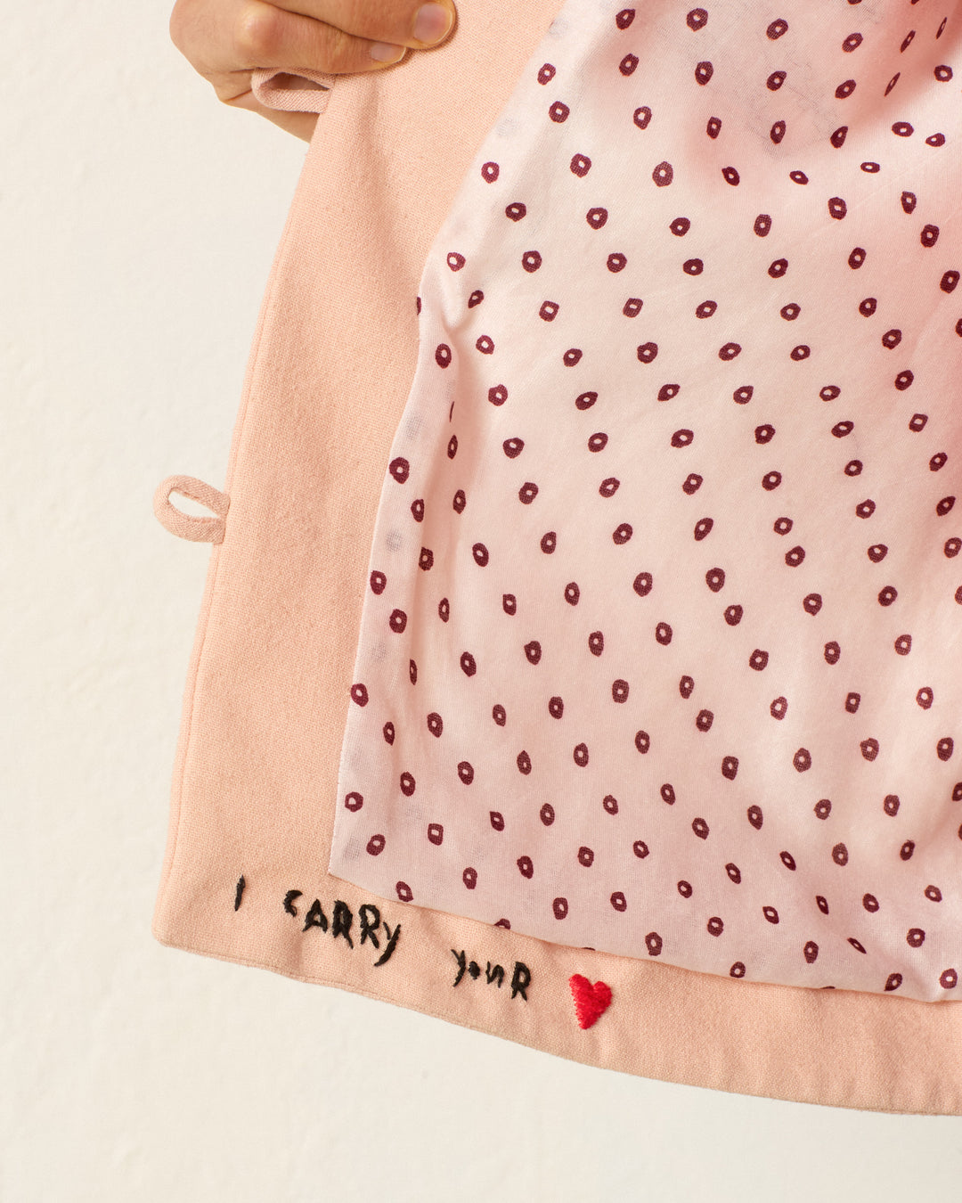A hand holds open the Jude Ruby Poppy Jacket, revealing its pink polka-dot lining and embroidered “I CARRY YOUR ❤” text on the inner hem.