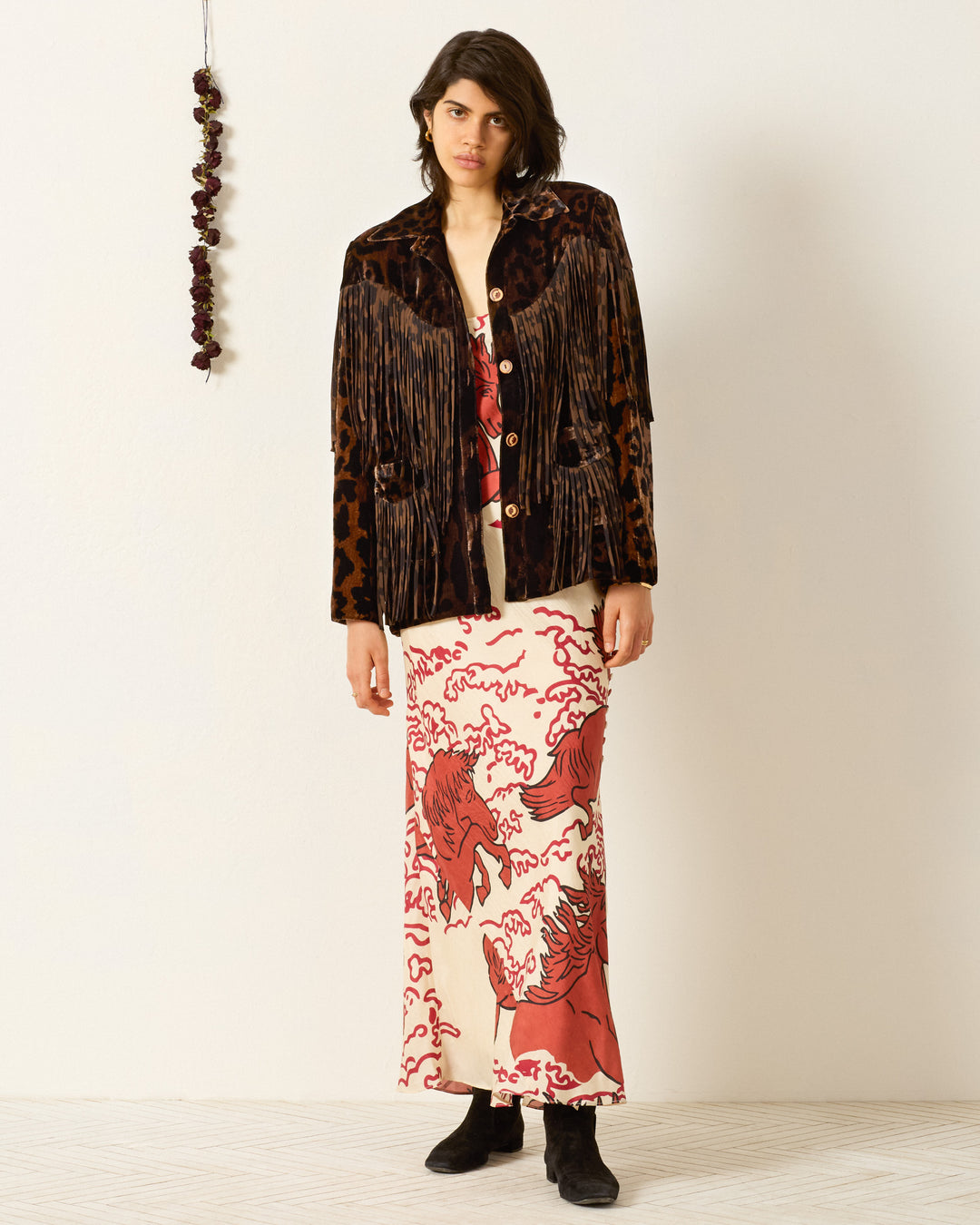 A person stands indoors wearing the Jude Leopard Velvet Jacket over a red and white printed dress with horse motifs, paired with black boots. A string of dried flowers hangs on the wall beside them.