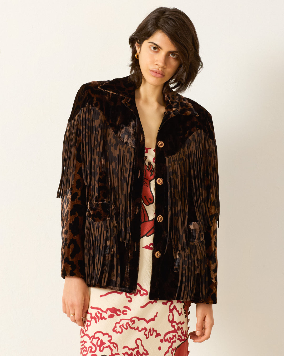 A woman wearing the Jude Leopard Velvet Jacket with Western-inspired details and silk fringe over a white and red patterned dress stands against a plain white background.