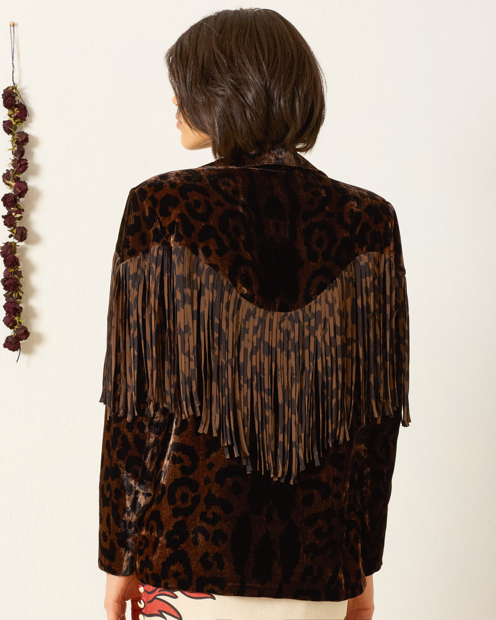 A person with short brown hair faces away, wearing the Jude Leopard Velvet Jacket—Western-inspired with a dark leopard print and brown and black fringe on the back. Dried flowers hang on the wall to the left.