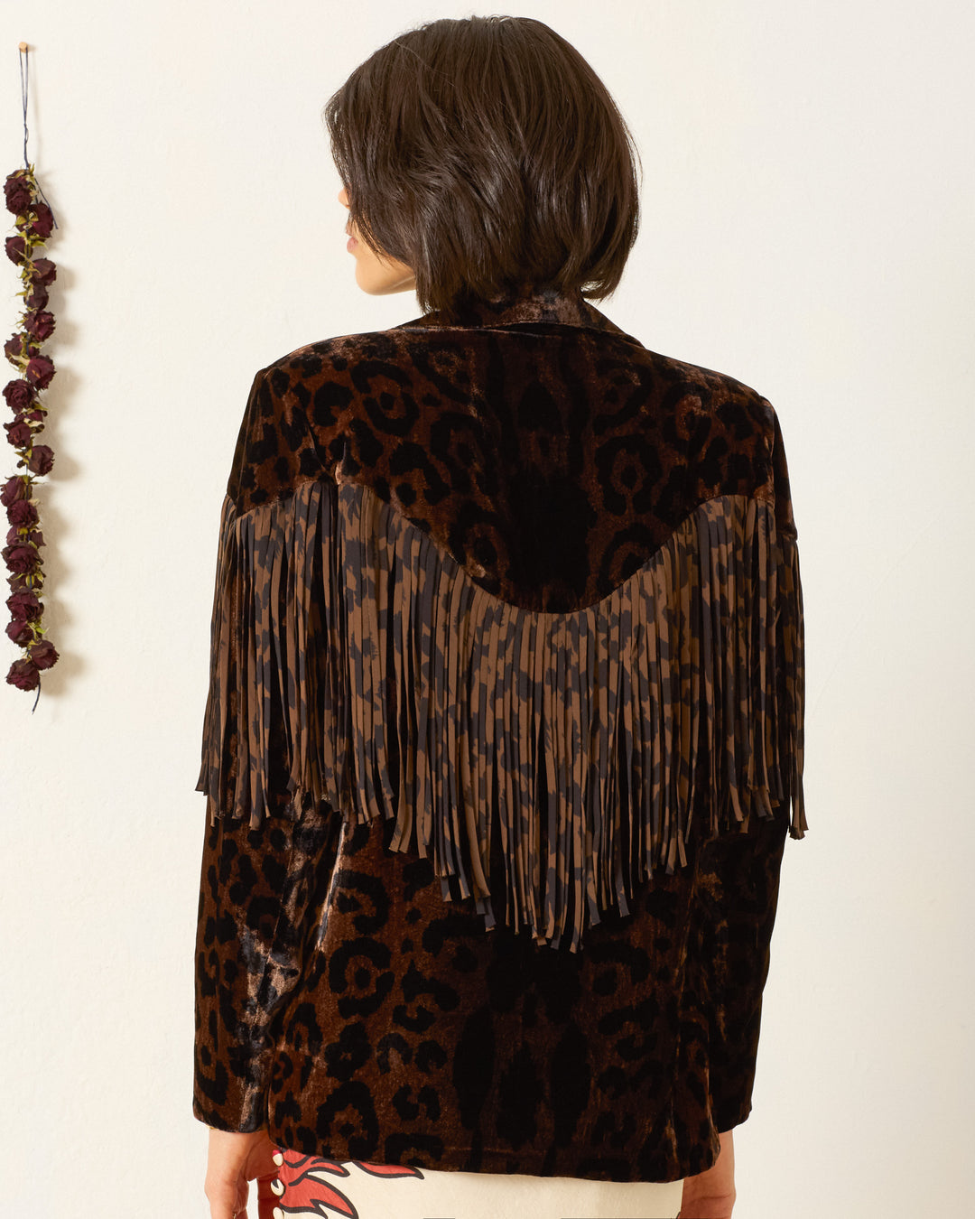 A person with short brown hair faces away, wearing the Jude Leopard Velvet Jacket—Western-inspired with a dark leopard print and brown and black fringe on the back. Dried flowers hang on the wall to the left.