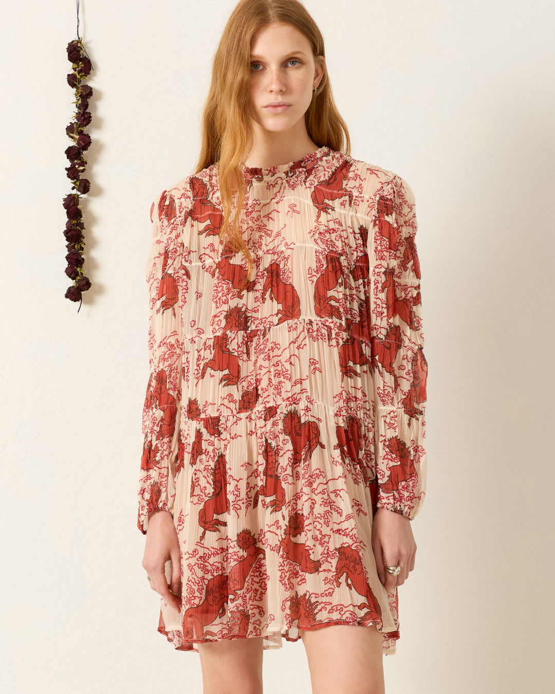 A woman stands indoors wearing the Juno Rouge Pegasus Dress, a long-sleeved cream dress with a red floral pattern and ruffle-trimmed neckline. Dried flowers hang on the wall beside her.