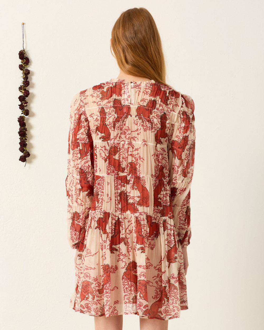 A woman with long red hair stands facing away, wearing the Juno Rouge Pegasus Dress, which features a semi-sheer red and cream pattern. Dried flowers hang on the white wall to her left.