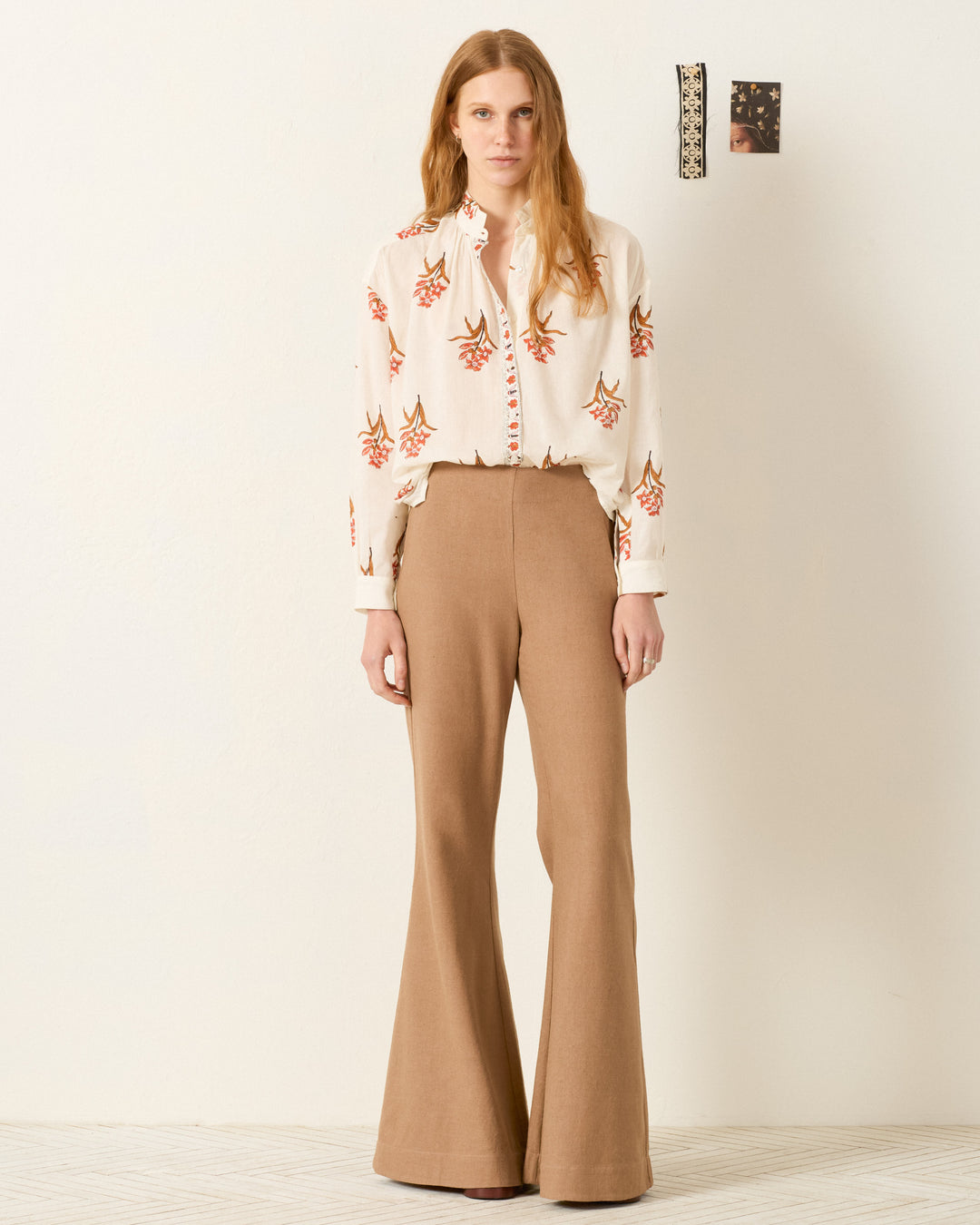 A woman stands indoors against a white wall, wearing the Kiki Persimmon Flower Shirt—a hand-block-printed cream blouse with red and brown floral patterns—paired with high-waisted, wide-leg tan pants for an elegant Edwardian look.