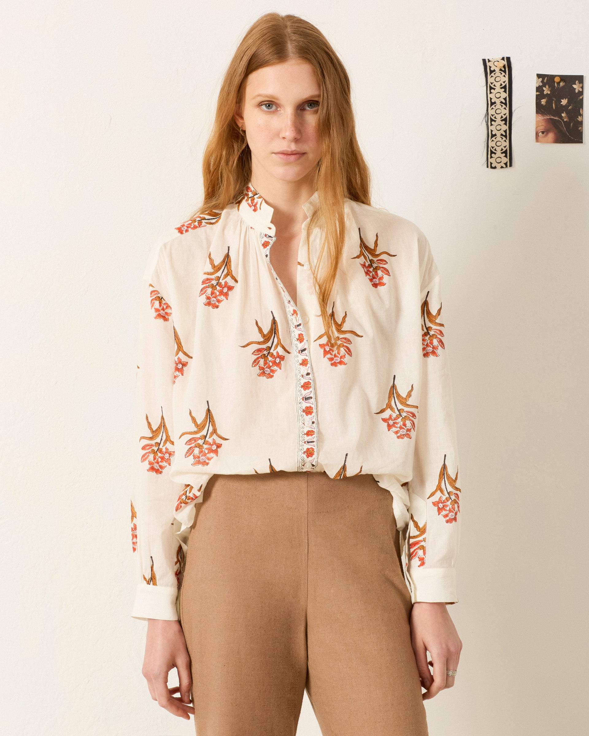 A woman with long blonde hair wears the Kiki Persimmon Flower Shirt and tan pants, standing against a plain off-white wall with two small art pieces in the background.