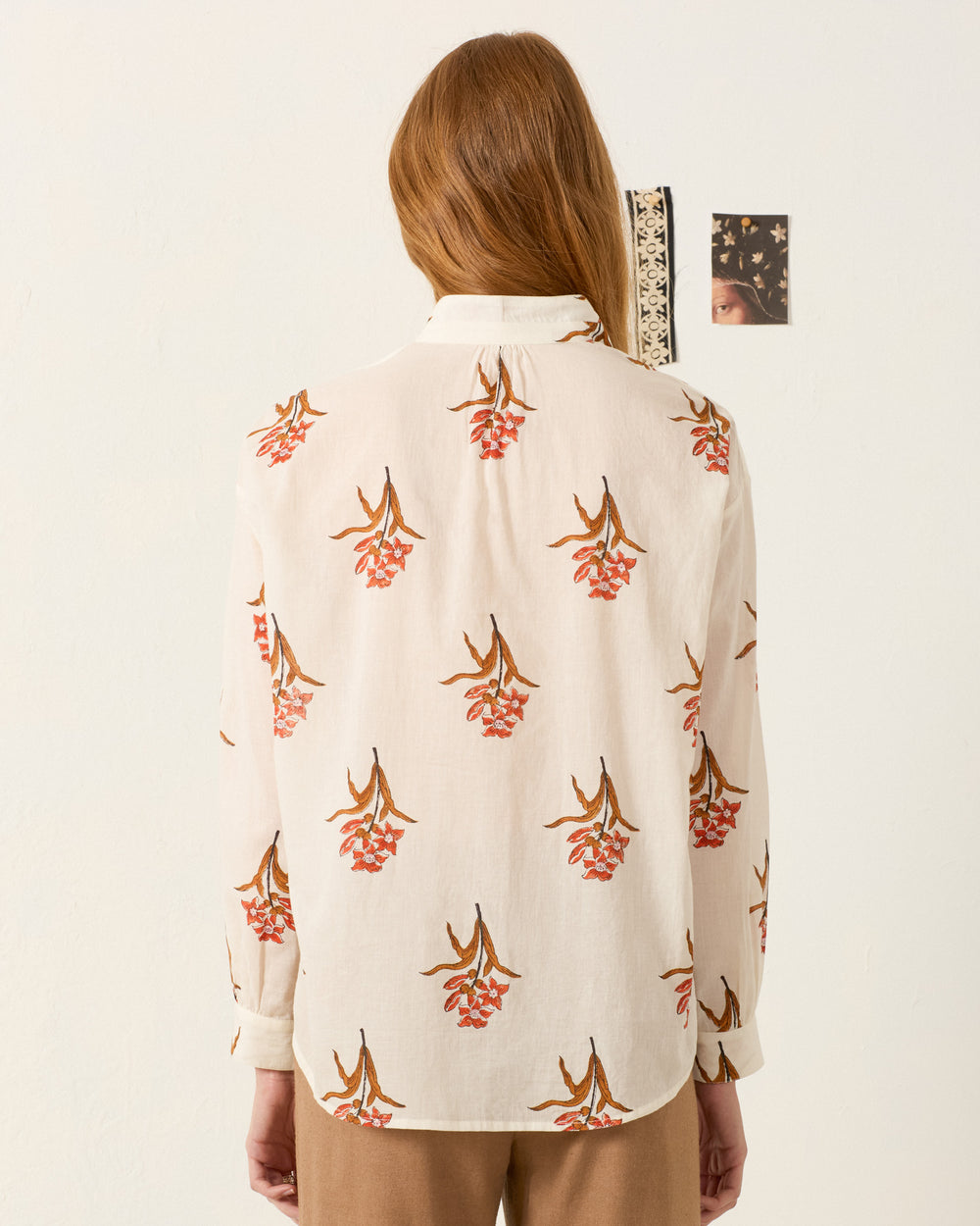 A person with long hair, seen from behind, wears the Kiki Persimmon Flower Shirt—hand-block-printed cotton voile with a red and brown floral pattern—standing before a wall decorated with small art pieces.