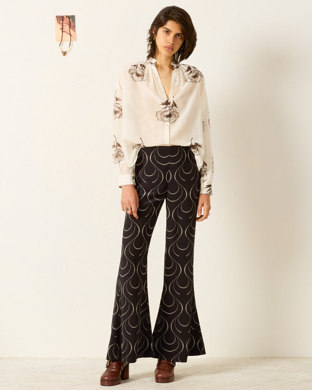 A person stands against a plain wall wearing the Kiki Obsidian Poppy Shirt, a hand-block-printed cream floral blouse with an Edwardian silhouette, paired with black patterned flared pants and brown platform shoes. A small photo is taped above their left shoulder.