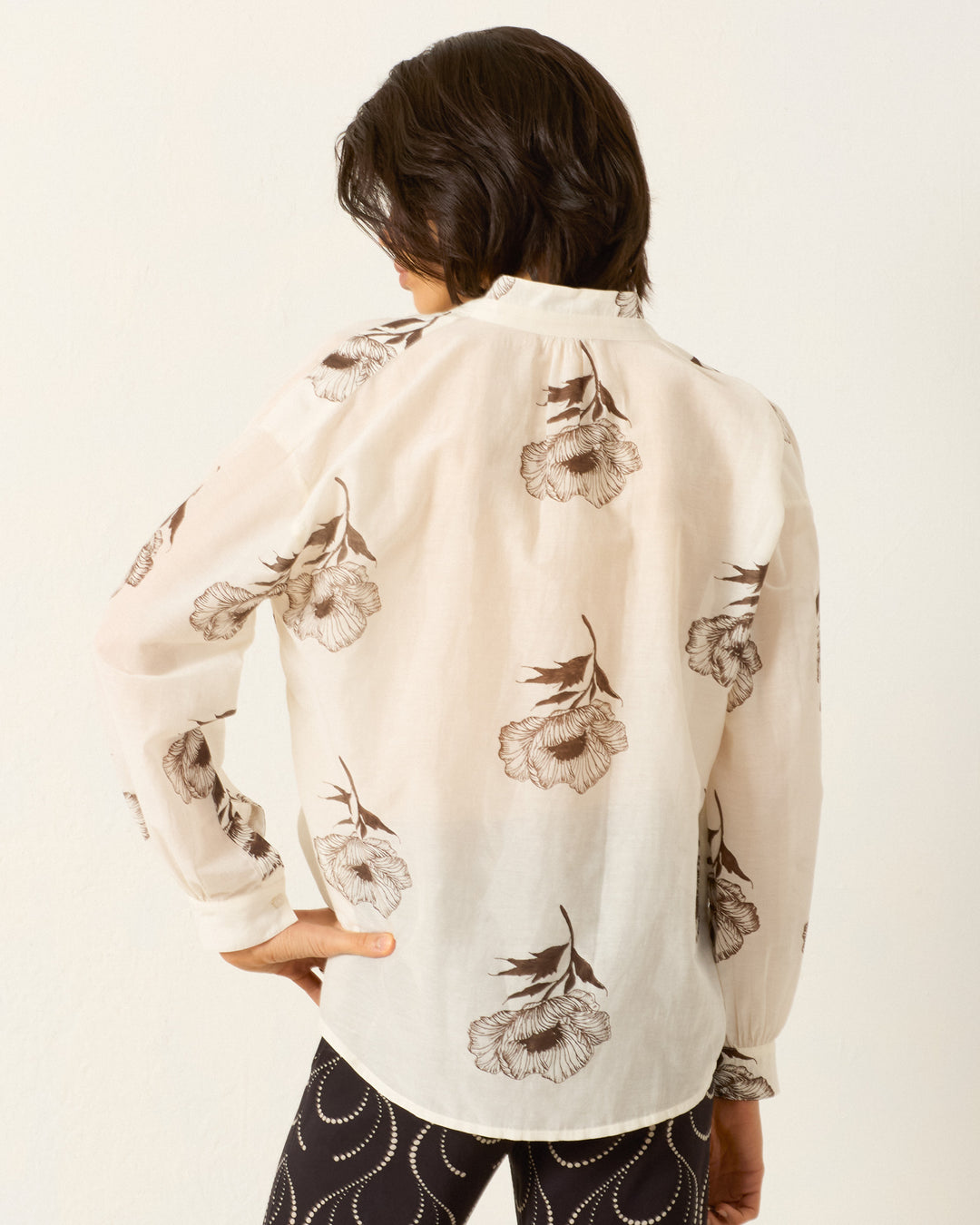 A person faces away, wearing the Kiki Obsidian Poppy Shirt—a sheer cotton-silk blend with a brown floral print—paired with dark pants featuring a white wavy pattern.