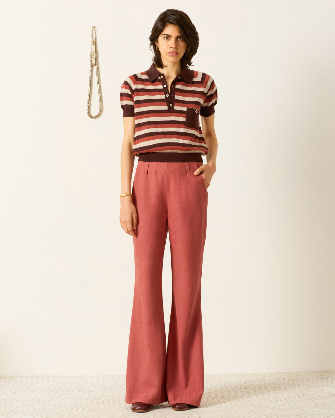 Indoors, someone wears the Lessi Sandstone Stripe Top with high-waisted, wide-leg red trousers and brown shoes. A rope is visible on the wall in the background.