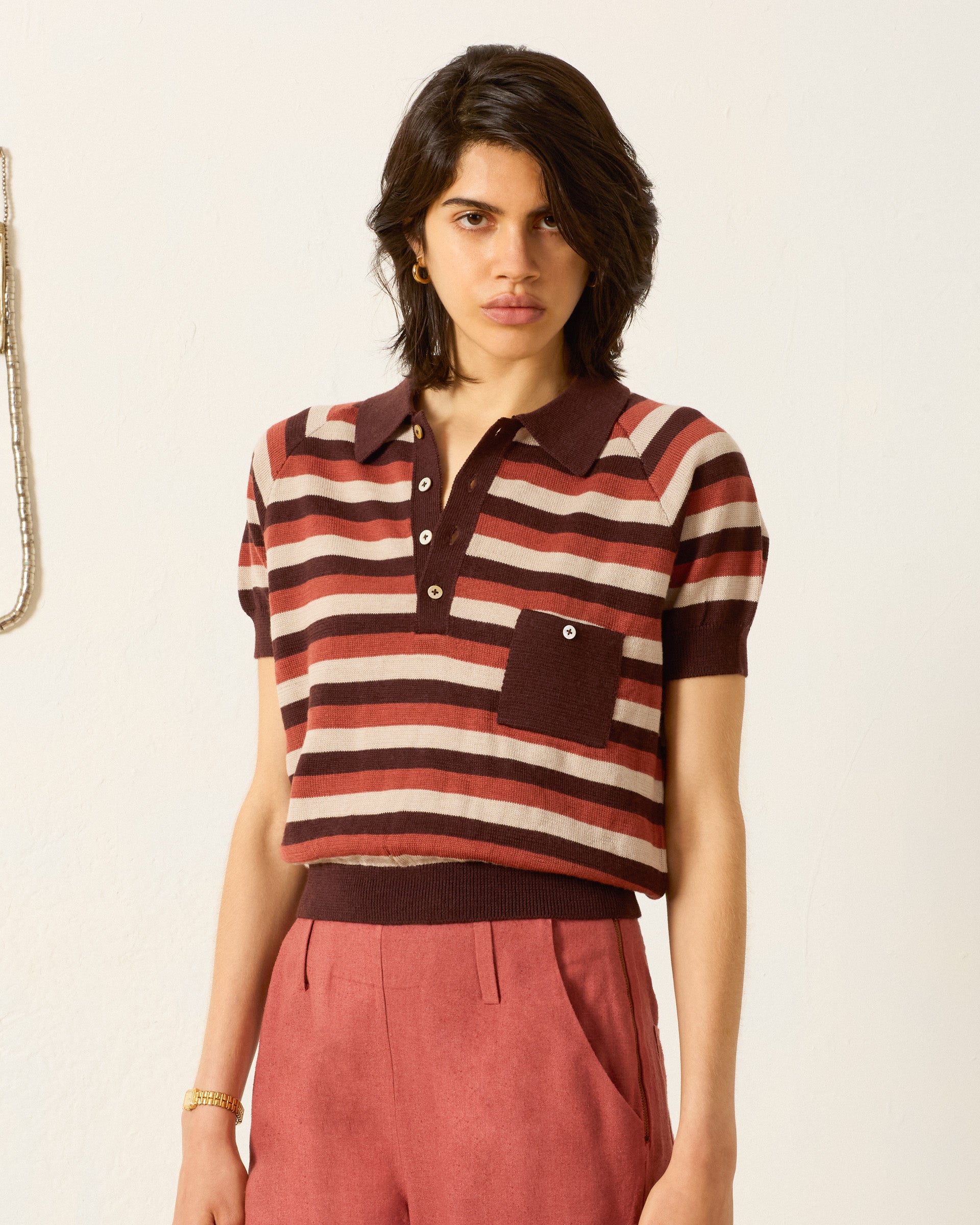 Person wearing the Lessi Sandstone Stripe Top, a short-sleeved maroon and beige striped polo sweater with a chest pocket, paired with high-waisted rust-colored trousers against a plain light background.