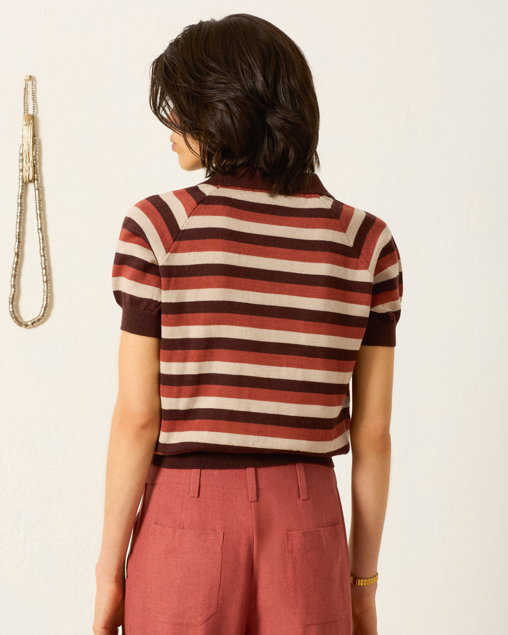 A person with short dark hair, facing away, wears the Lessi Sandstone Stripe Top—a short-sleeved, striped polo sweater in brown and beige—paired with rust-colored pants. A beaded rope is visible on the wall to the left.
