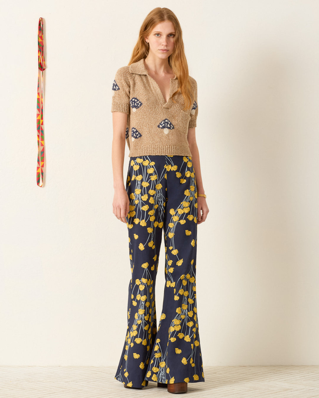 A woman stands indoors wearing the Lessi Mushroom Knit Top, a beige cropped polo sweater, paired with navy-blue flared pants featuring a yellow floral print. A patterned belt hangs on the wall beside her.