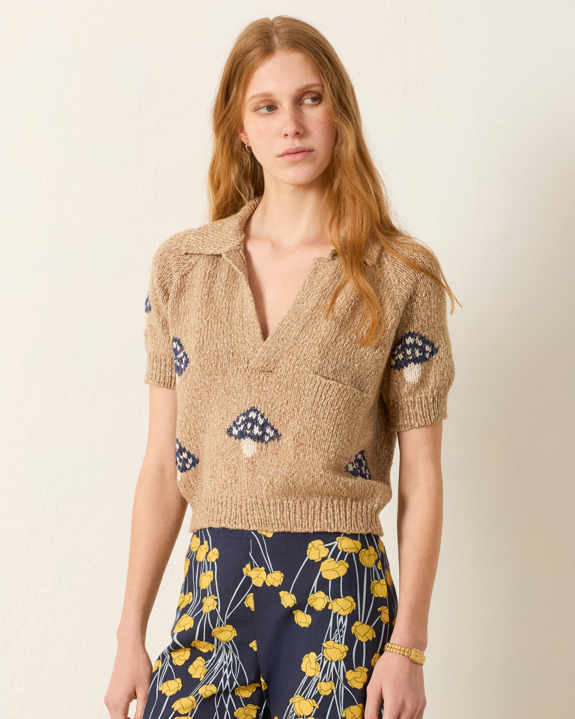 A woman with long light brown hair wears the Lessi Mushroom Knit Top, a beige short-sleeve polo sweater with blue patterns and a collar, styled with dark pants that have a yellow floral print.