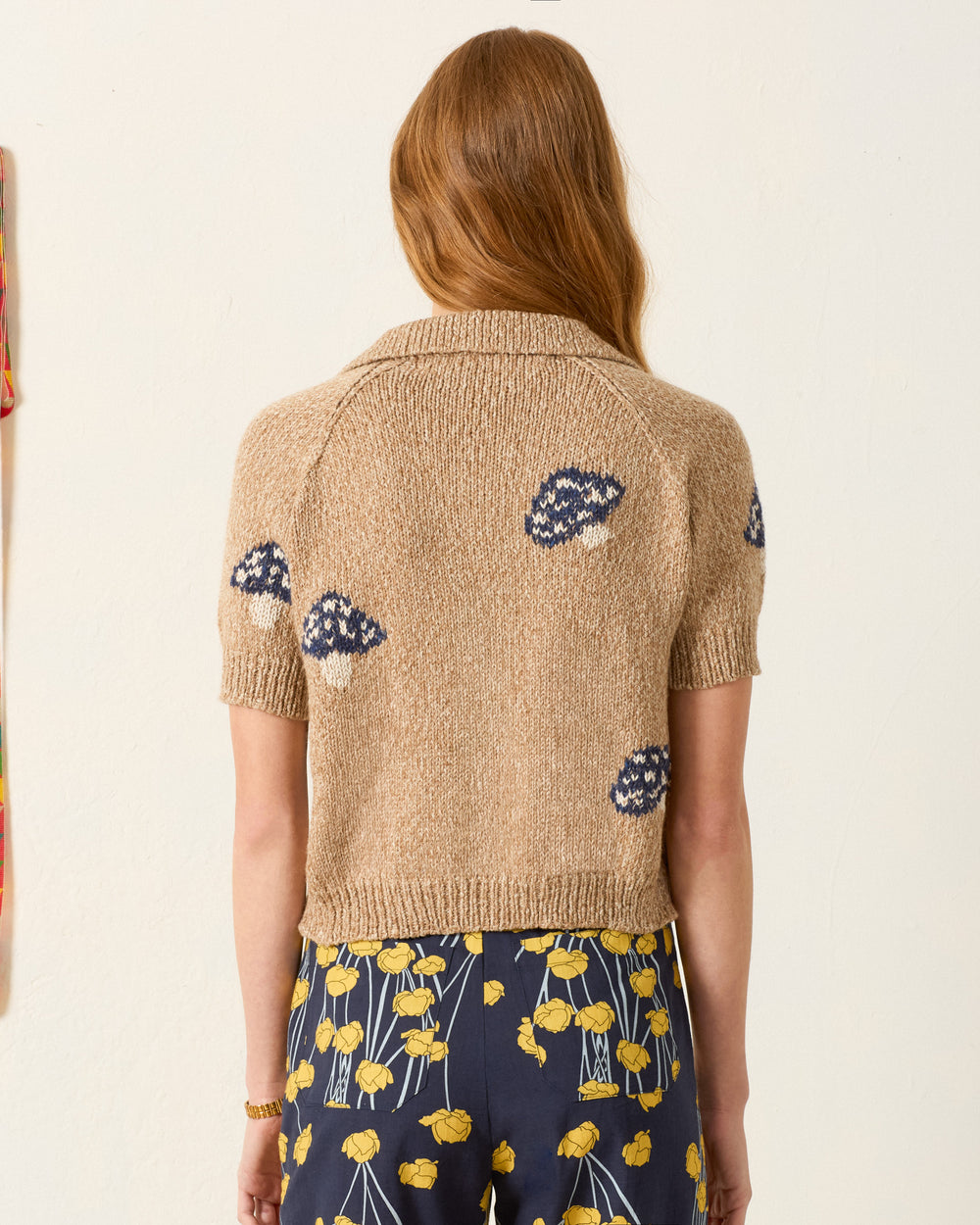 A woman with long reddish hair, seen from behind, wears the Lessi Mushroom Knit Top—a cropped baby alpaca sweater with blue patterns—paired with a navy skirt featuring a yellow floral print.