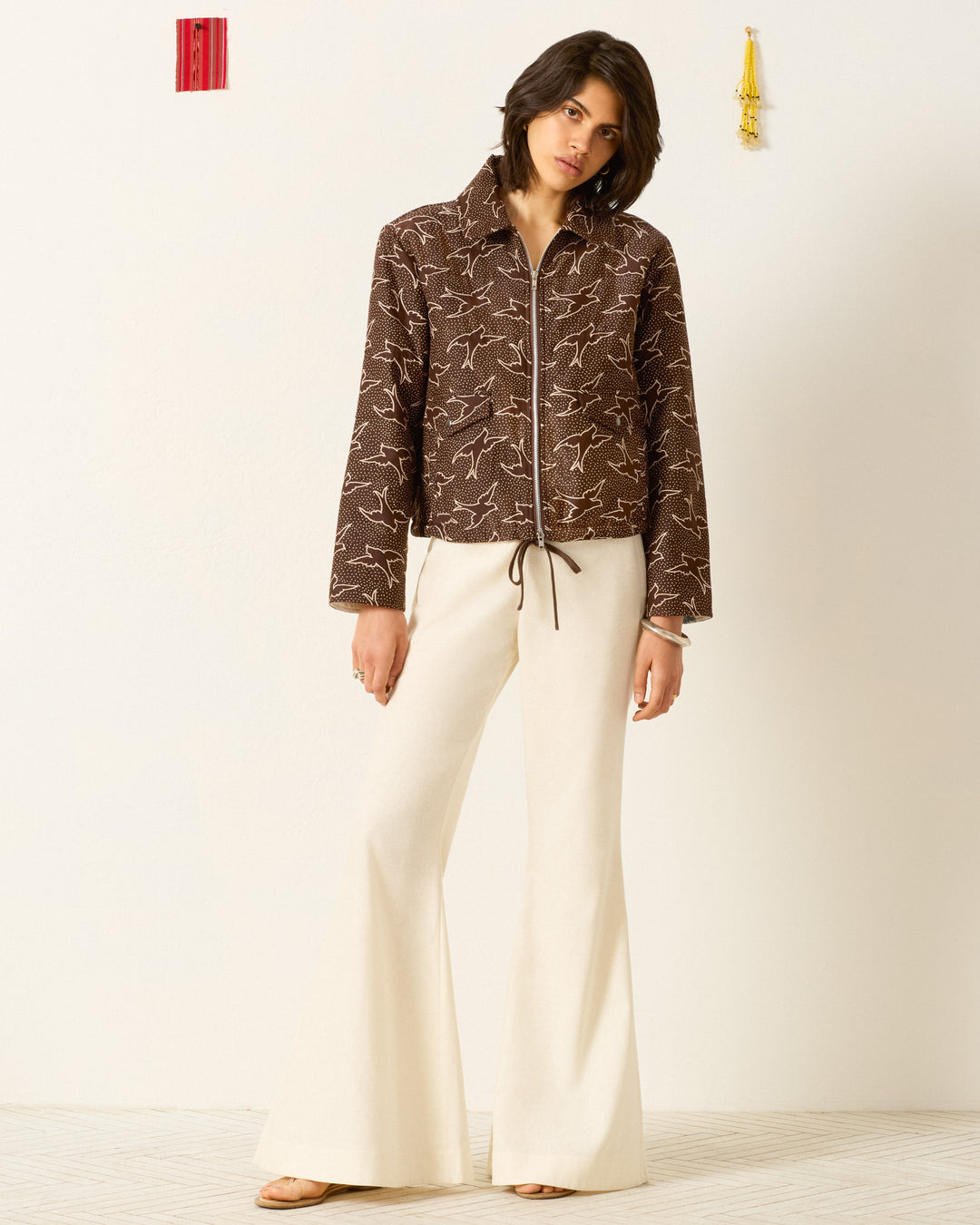 A woman stands against a plain wall wearing the Lex Clove Swallow Jacket, featuring a brown pattern with subtle swallow motifs, paired with cream flared pants and sandals.