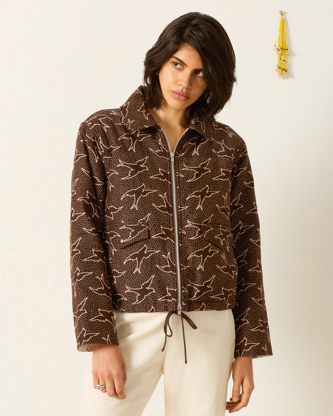 Someone stands against a plain white wall with a yellow hanging decoration, wearing cream pants and the Lex Clove Swallow Jacket—a brown workwear zip jacket featuring a swallow motif.