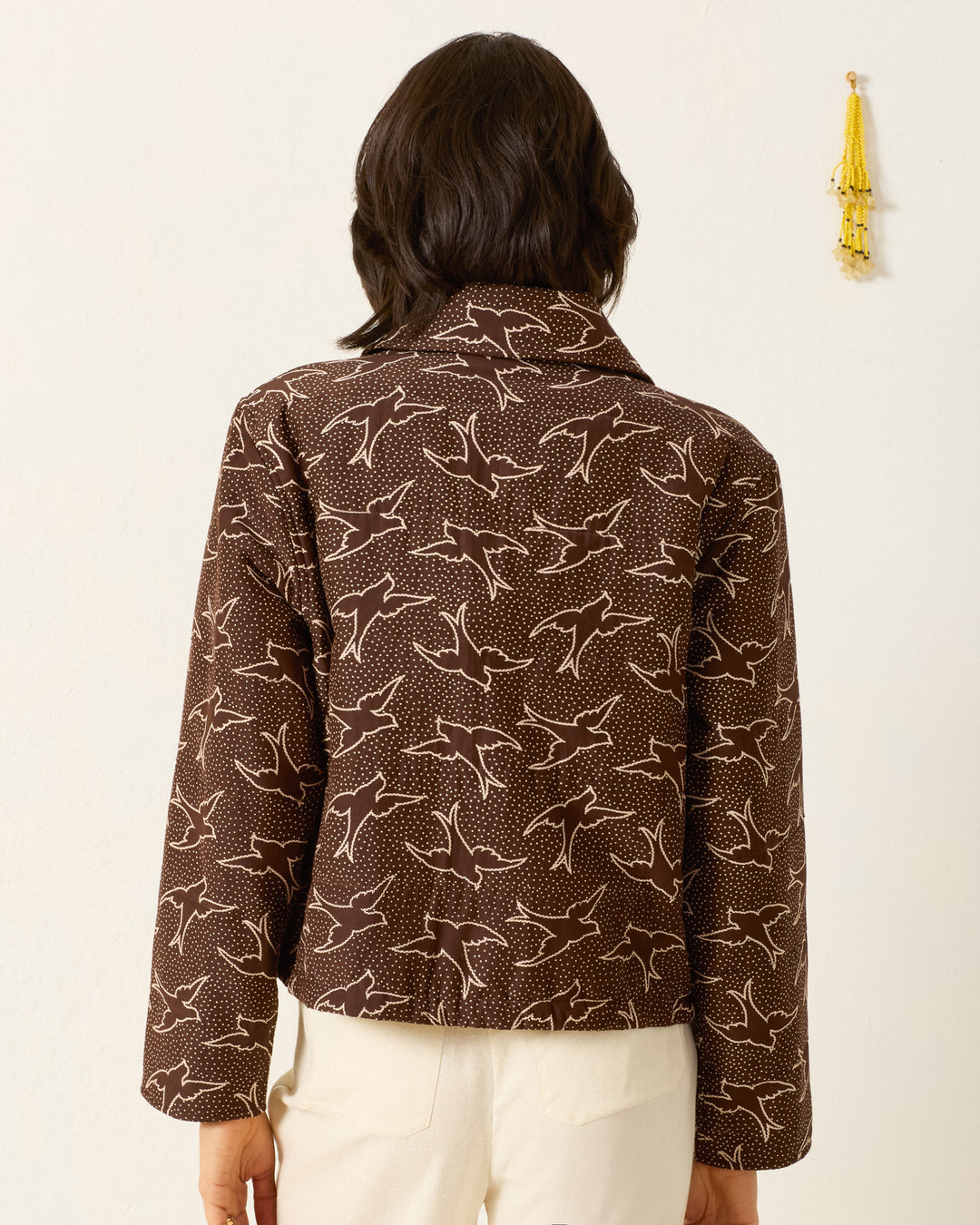 A person with dark hair is shown from the back, wearing the brown Lex Clove Swallow Jacket with a white swallow motif and cream pants. A small yellow tassel decorates the wall.