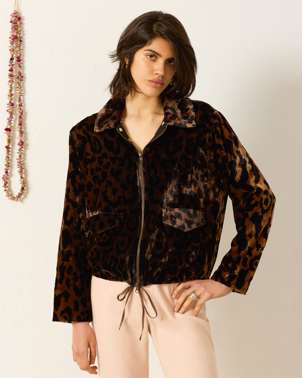 Wearing the Lex Leopard Velvet Jacket, featuring a brown and black leopard print, zip front, and drawstring hem, paired with light beige pants against a white wall for a chic twist on classic workwear.