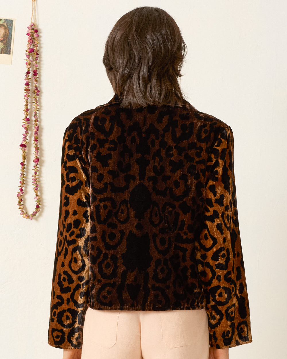 A person with shoulder-length brown hair is seen from behind, wearing the Lex Leopard Velvet Jacket and light-colored pants, standing indoors near a wall decorated with hanging ornaments.