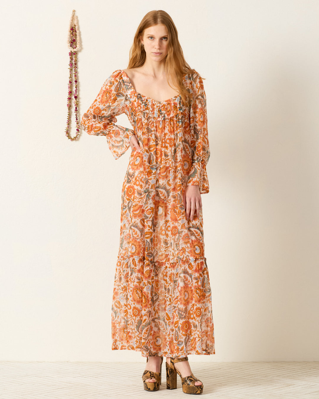 Indoors, a woman wears the Lilium Topaz Flower Dress—a pleated empire waist maxi with a vibrant floral motif and long sleeves—paired with platform heels. A beaded necklace hangs on the wall beside her.