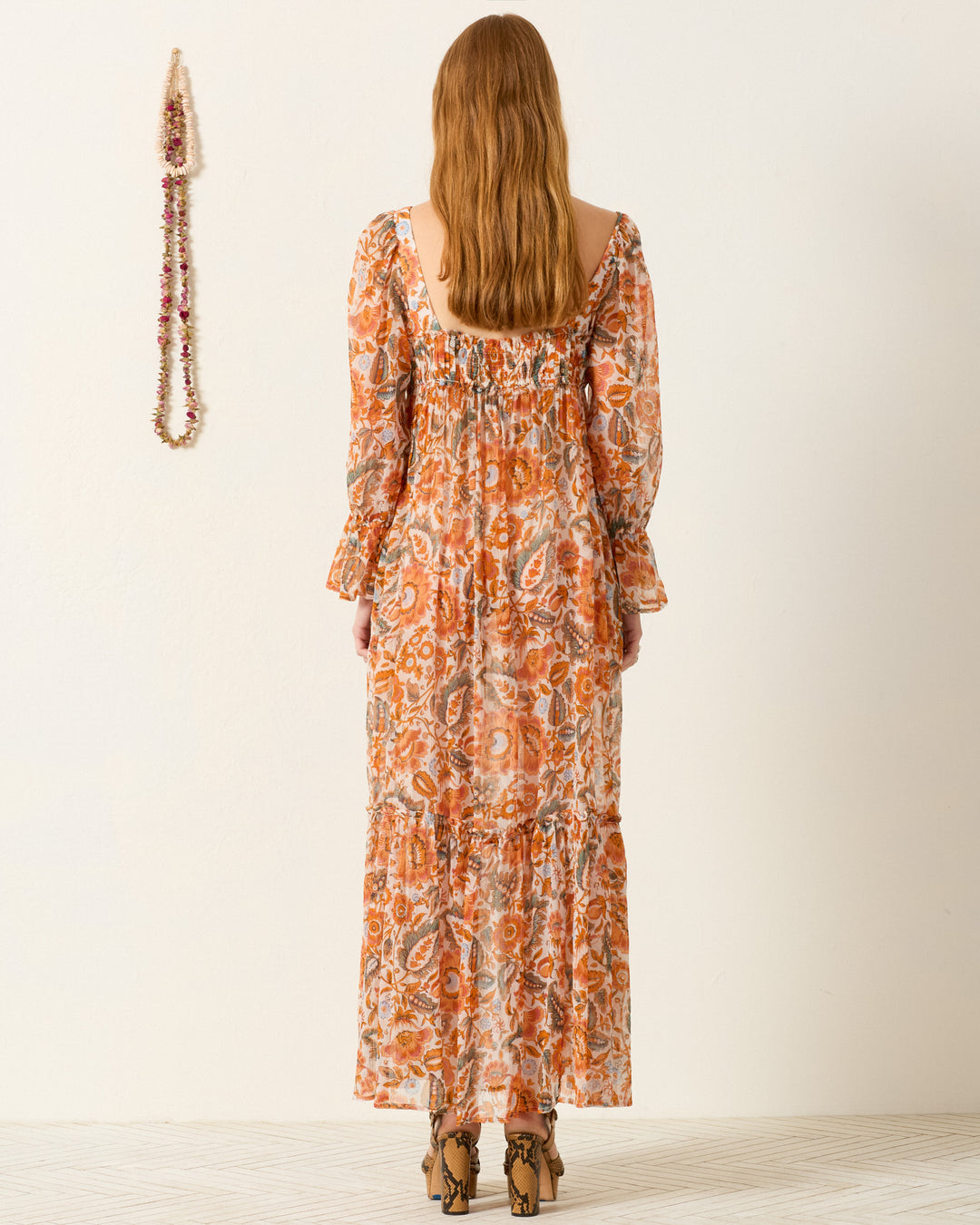 A woman with her back to the camera wears the Lilium Topaz Flower Dress—an orange and cream maxi with a floral print, long sleeves, and a tiered skirt—against a plain background.