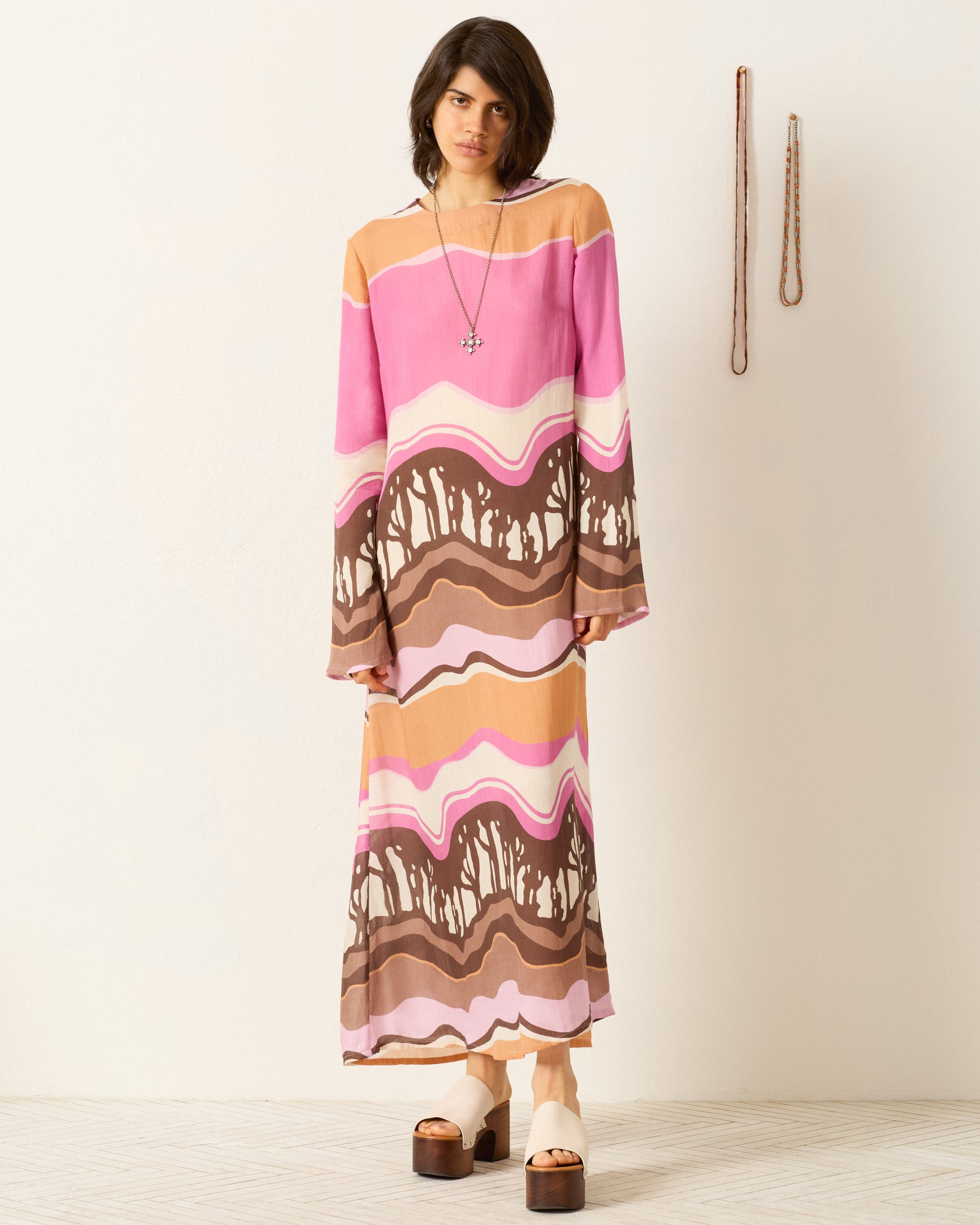 A person stands indoors wearing the Niall Bloomshade Dress, featuring pink, brown, and beige abstract wavy stripes, paired with platform sandals and a pendant necklace. Two belts are hanging on the wall beside them.