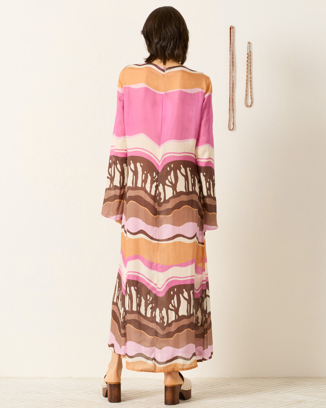 A person stands facing a wall, wearing the Niall Bloomshade Dress, a colorful maxi with an abstract pink, brown, and orange print. Two necklaces hang on the wall to the right.
