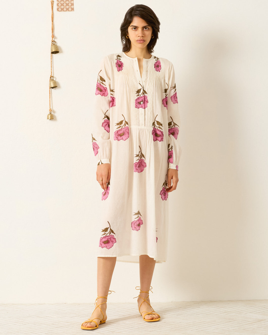 Indoors, a person wears the Nyxe Fuchsia Poppy Dress—a white cotton voile shirtdress with pink hand-block-printed florals—paired with yellow lace-up sandals.