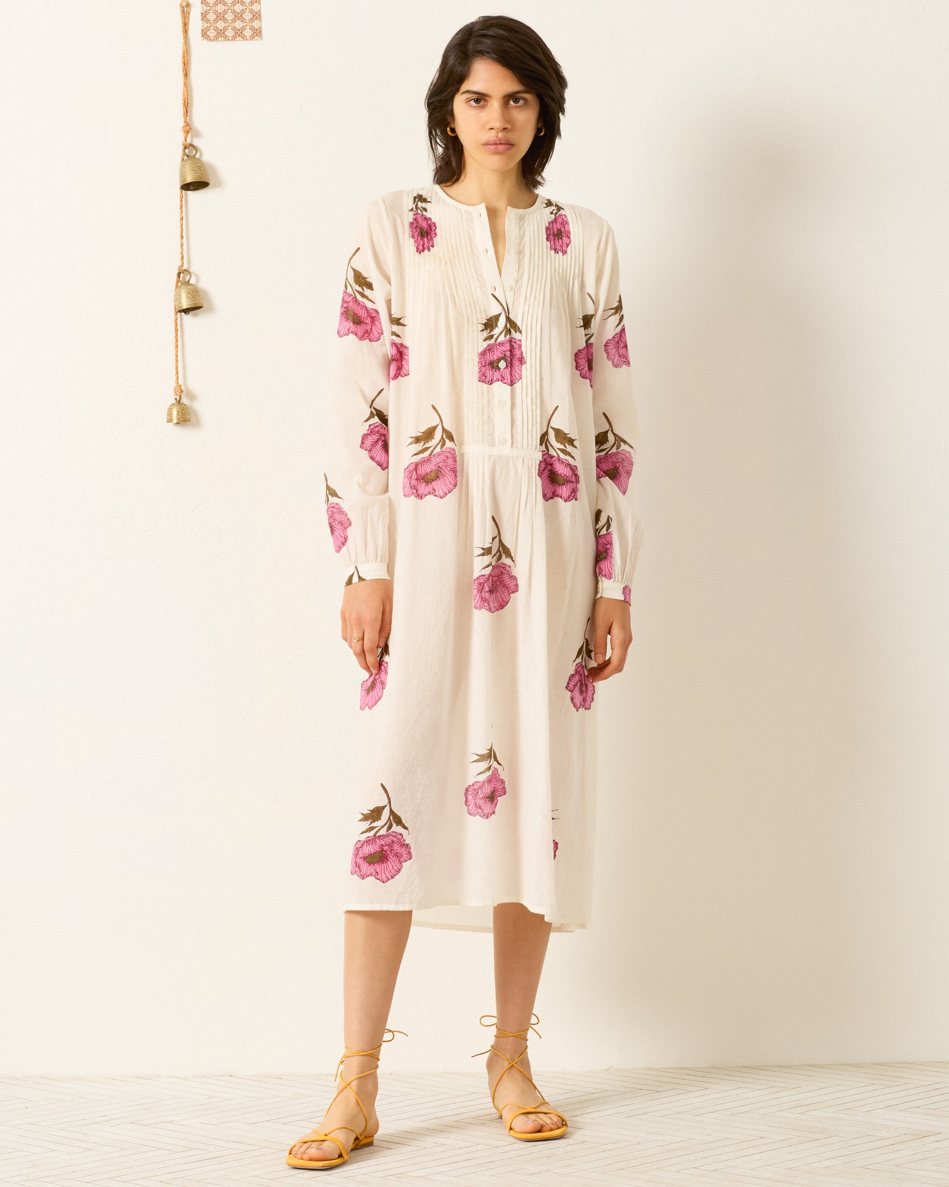 Indoors, a person wears the Nyxe Fuchsia Poppy Dress—a white cotton voile shirtdress with pink hand-block-printed florals—paired with yellow lace-up sandals.