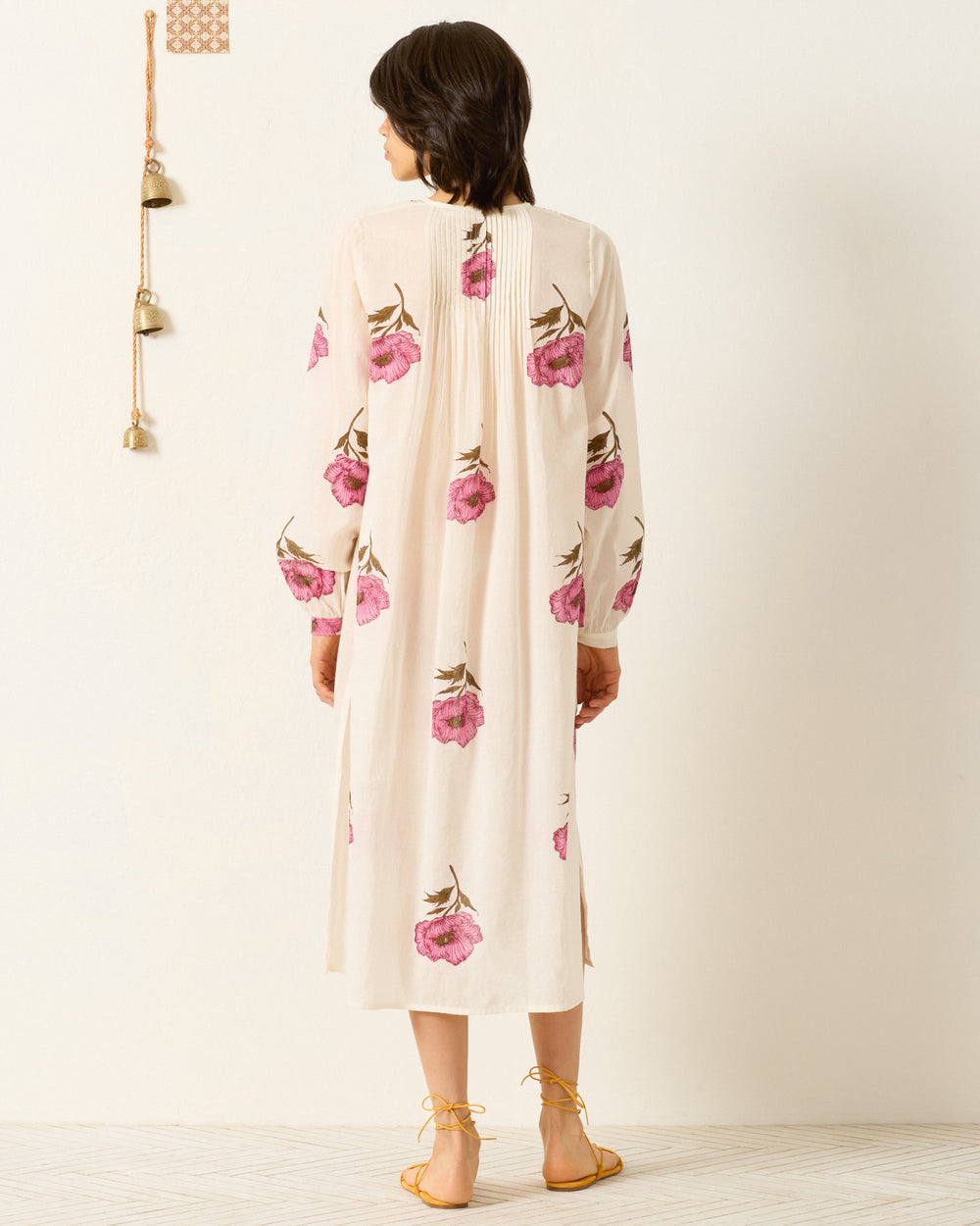 A woman stands barefoot facing away, wearing the Nyxe Fuchsia Poppy Dress— a long cream cotton voile shirt dress with pink hand-block-printed poppy motifs and long sleeves—set against a plain backdrop with bells hanging on the wall.