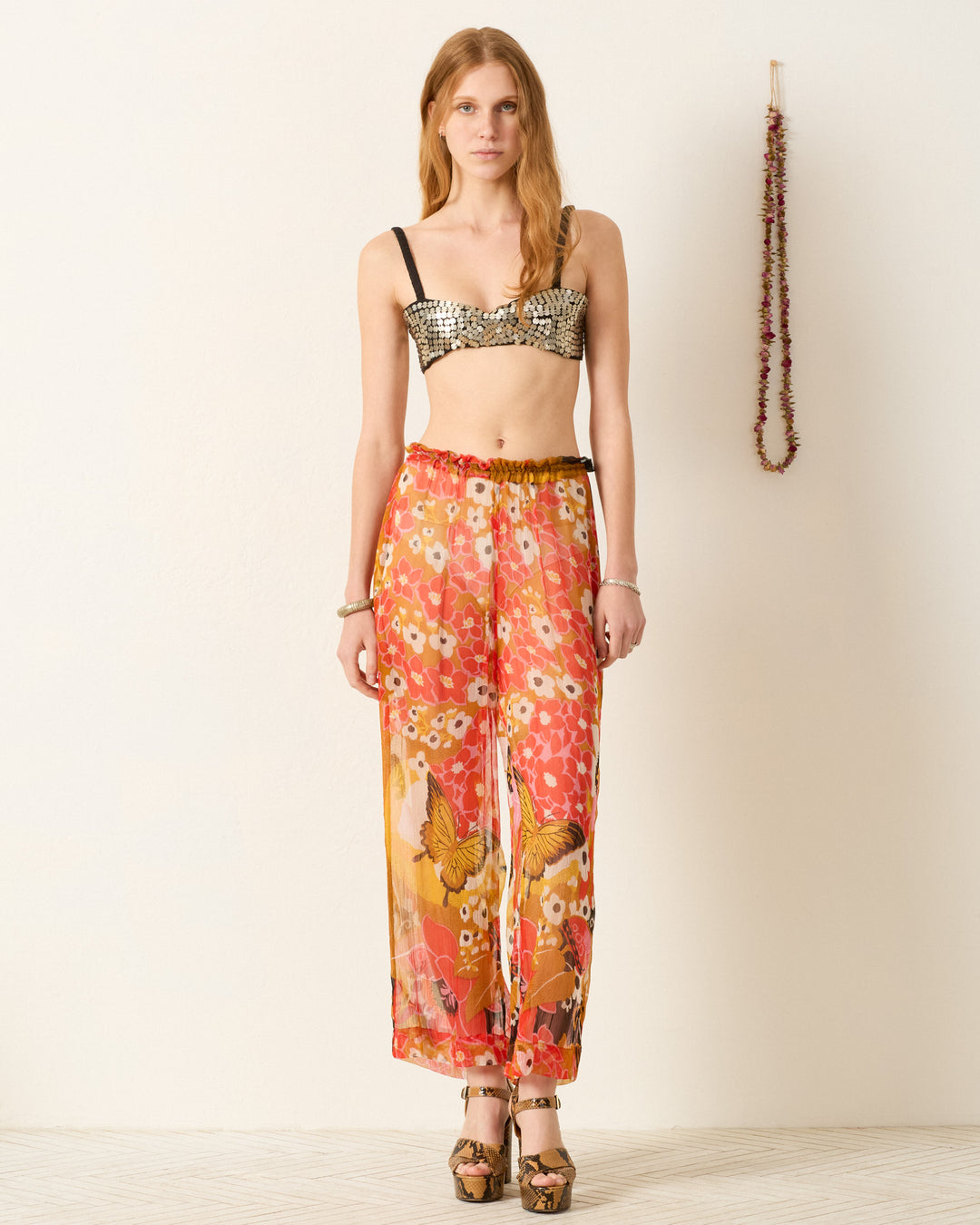 A woman stands indoors wearing the Otto Marigold Meadow Pant with an elasticated waist, a sequined bralette, and platform sandals; a garland decorates the wall behind her.