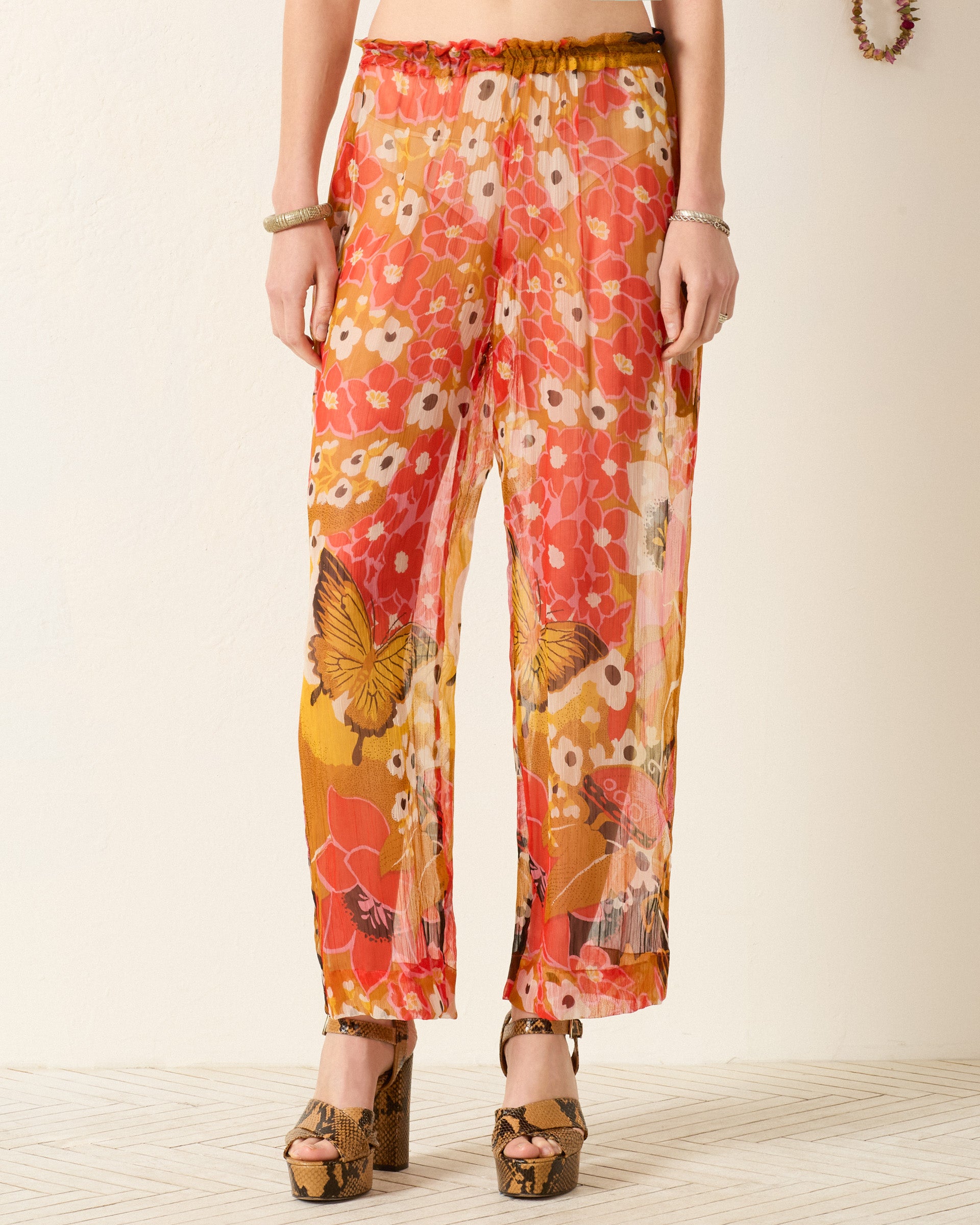 A person wears the Otto Marigold Meadow Pant—sheer, floral with butterfly prints and an elasticated waist—styled with platform sandals against a light-colored wall.