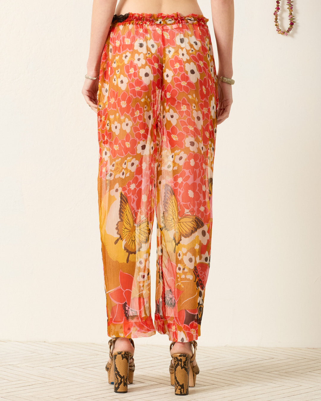 Person wearing the Otto Marigold Meadow Pant, featuring a flowy pajama-style silhouette, elasticated waist, and red, orange, and yellow butterfly pattern, seen from the back while standing on a light-colored floor.