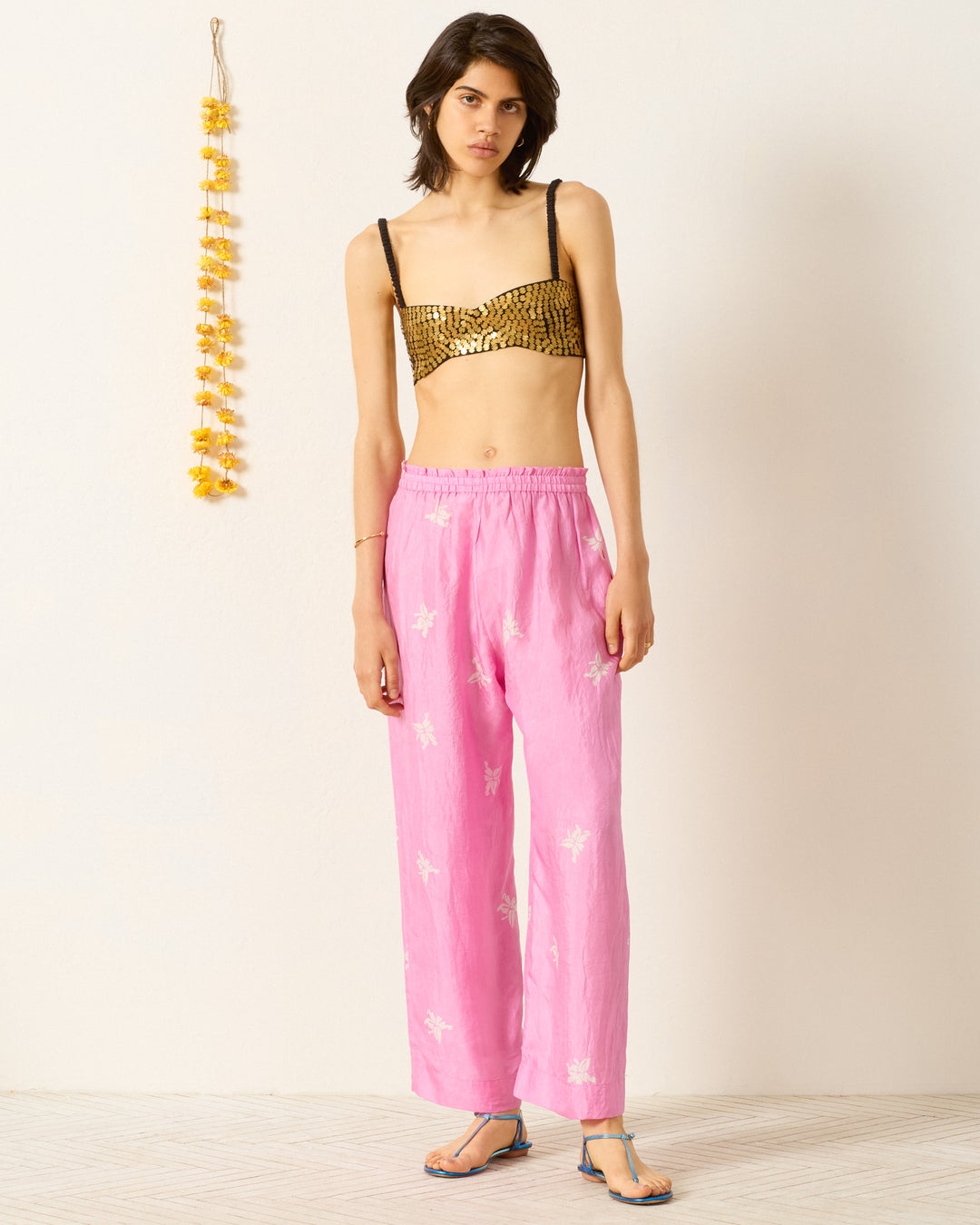 A person stands against a plain wall wearing a gold sequin bandeau top, Otto Pink Jasmine Pant with white patterns, and sandals. A yellow flower garland hangs on the left.
