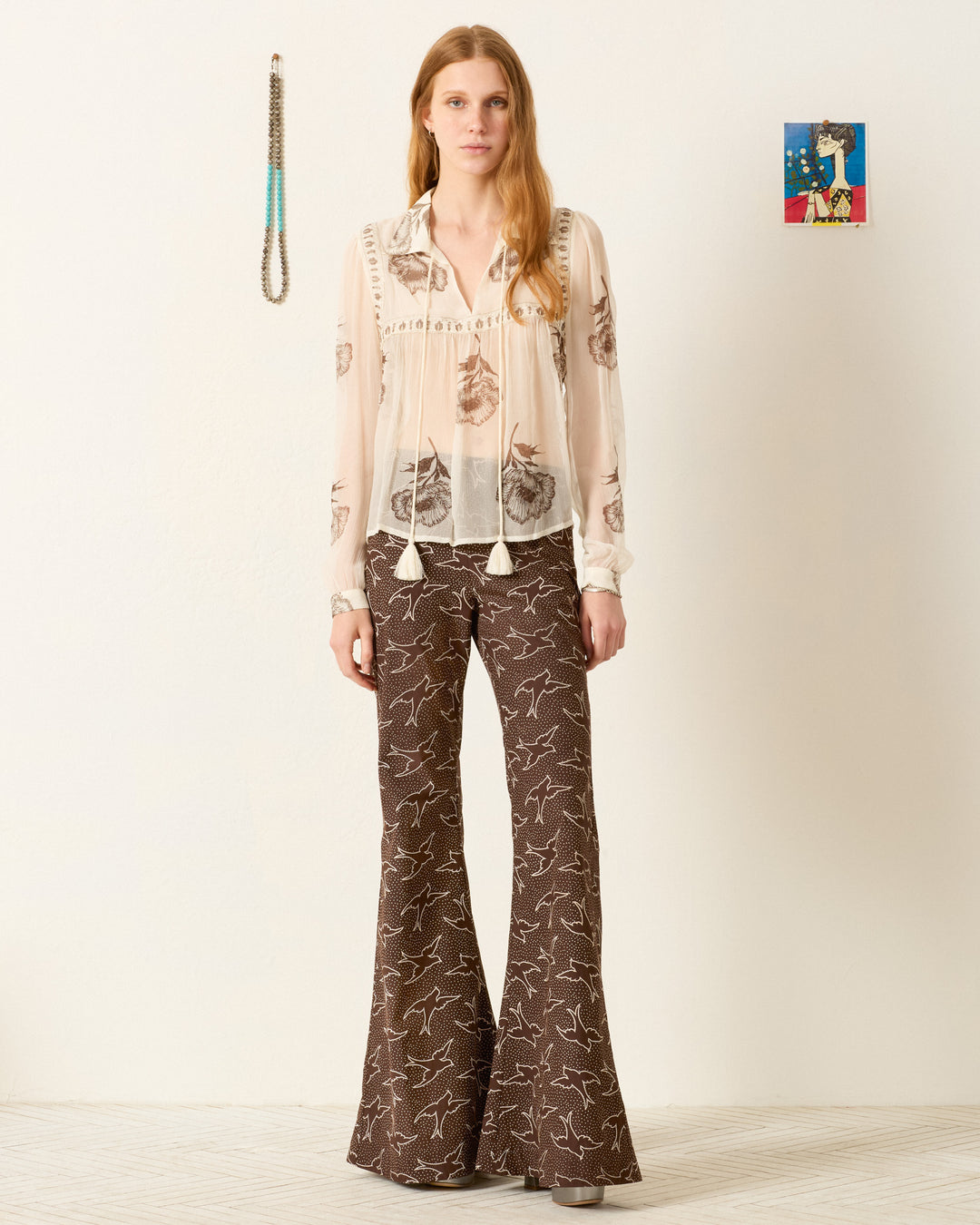 A person stands indoors wearing the Pascal Obsidian Poppy Top, a semi-sheer hand-block-printed cupro-chiffon piece, paired with brown flared trousers featuring a white bird print; art and a necklace hang on the wall behind.
