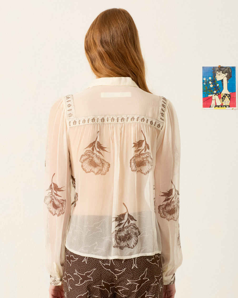 A woman with long hair, seen from behind, wears the Pascal Obsidian Poppy Top and patterned brown pants, standing against a white wall with a small colorful artwork.