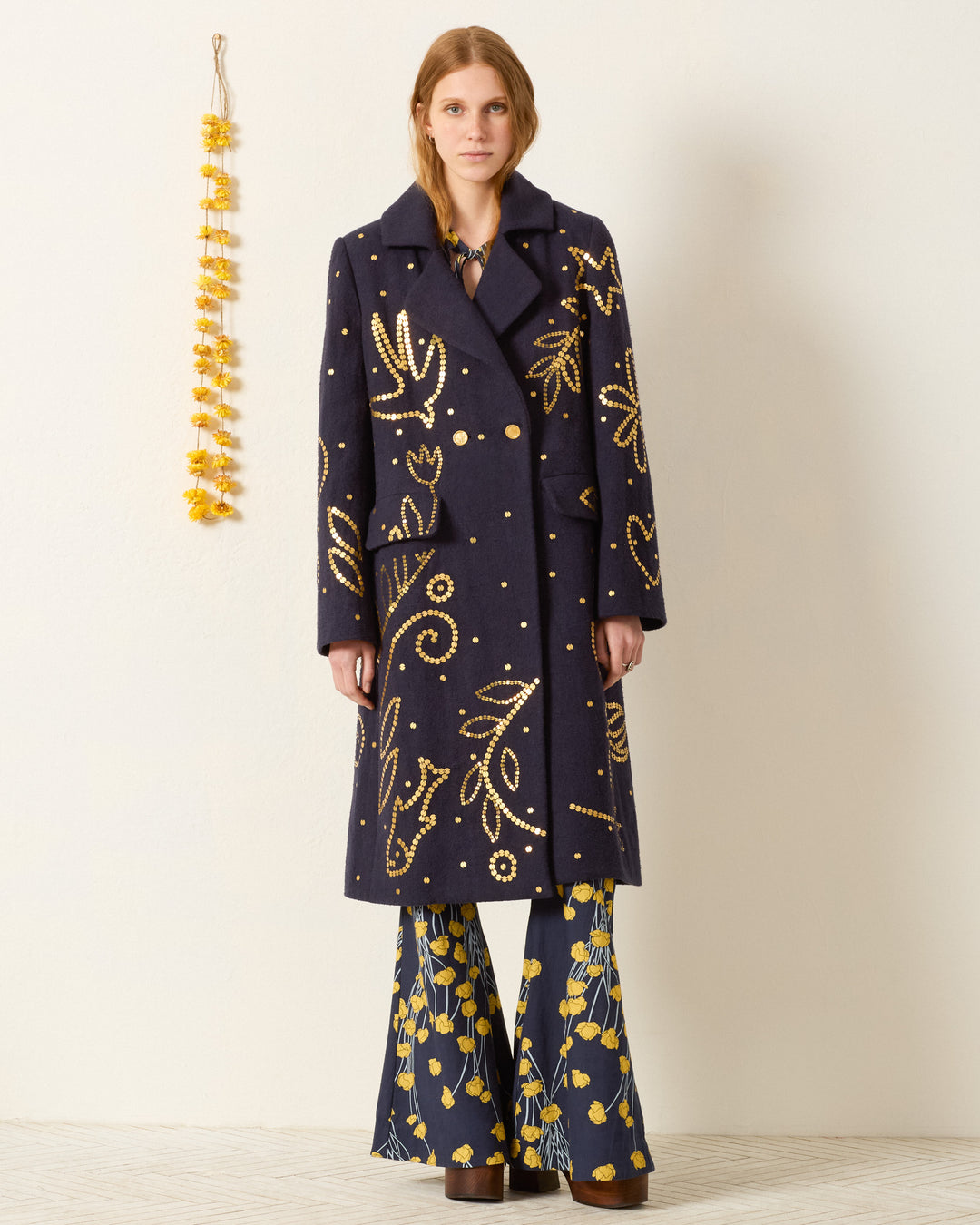 A woman stands against a plain wall wearing the Petunia Midnight Coin Coat in navy blue with yellow patterns and flared blue pants with yellow designs. A yellow garland hangs on the wall beside her.