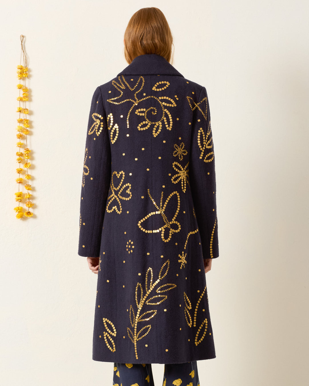 A person with long hair, seen from behind, wears the hand-embellished Petunia Midnight Coin Coat featuring yellow floral and leaf patterns. A yellow garland hangs on the wall to the left.